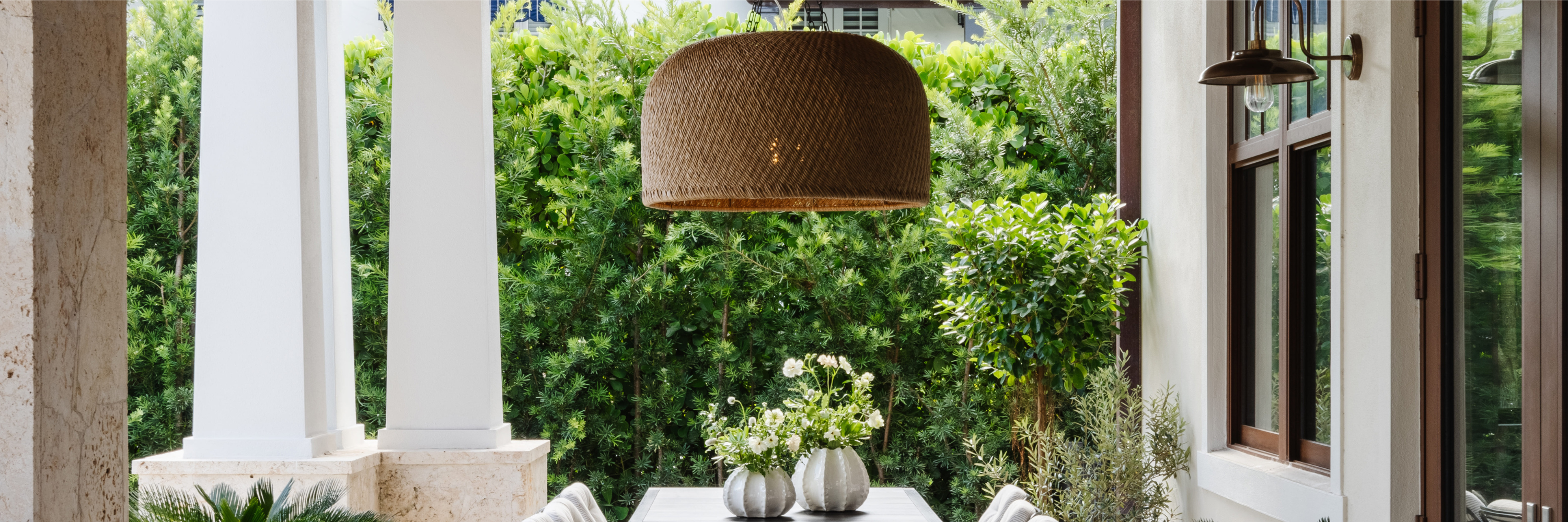 Outdoor dining area with a large woven pendant light hanging above a rectangular table set with neutral vases and greenery. Surrounded by lush hedges and framed by stone columns and wood-trimmed windows, the space feels warm and inviting.
