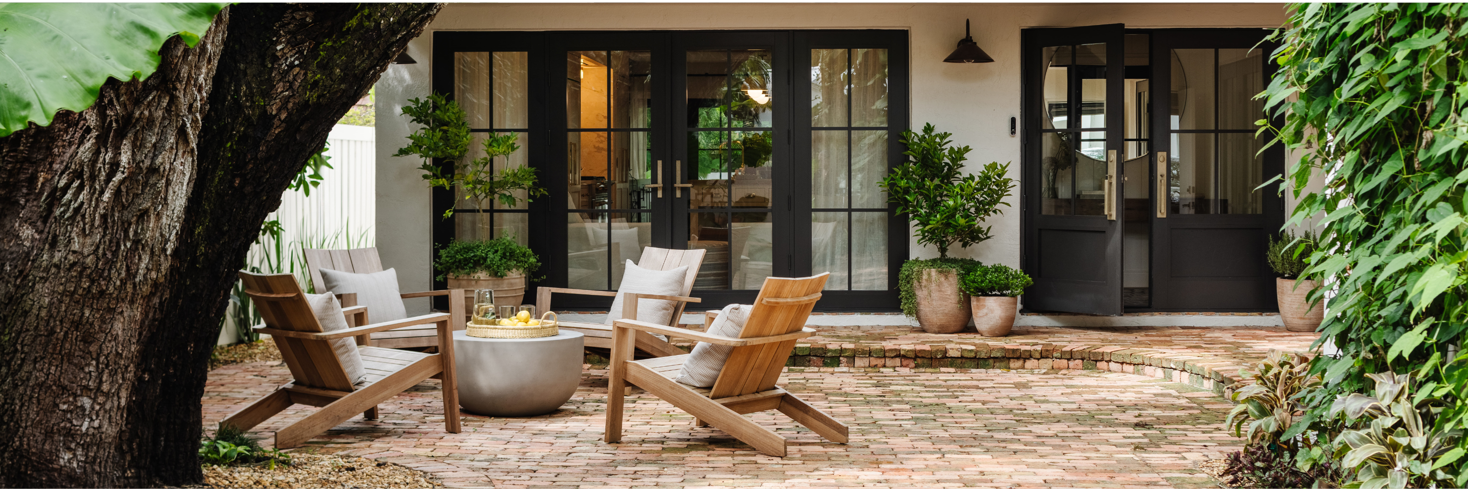 Outdoor patio with four wooden lounge chairs arranged around a round stone table, soft cushions, layered greenery in planters, brick flooring, and black French doors opening to the interior of a home designed by Calimia Home.
