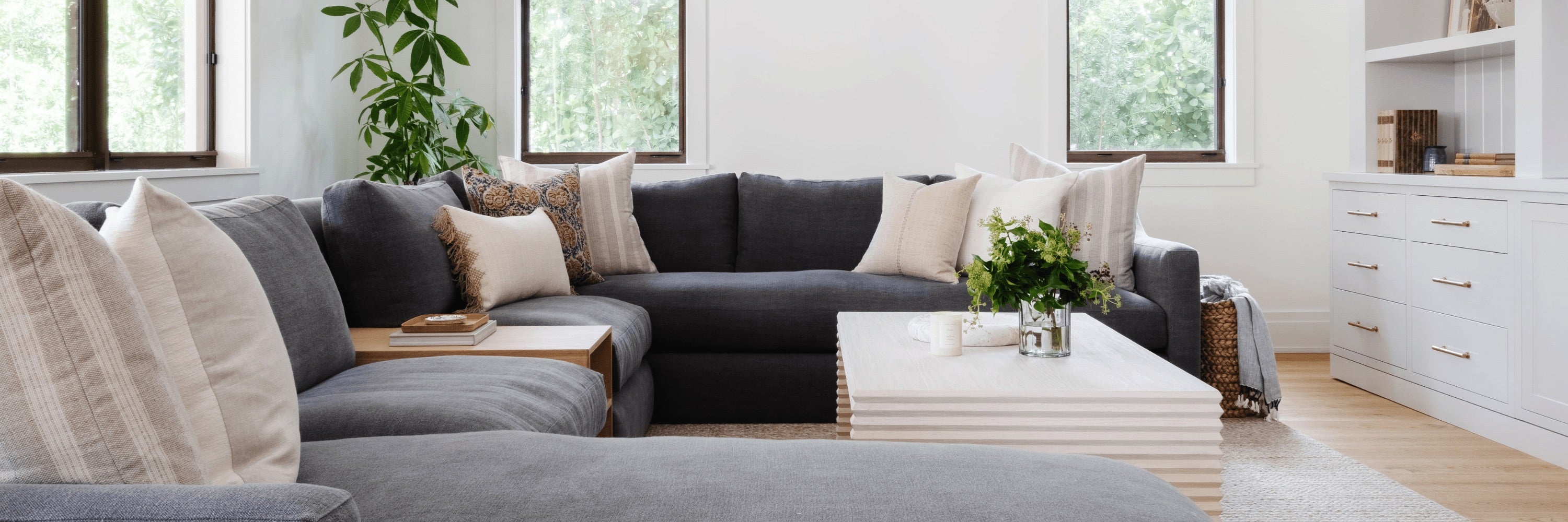 Upholstered sectional sofa with layered linen pillows, throws, and soft ottomans in a sunlit living room, from the Calimia Home furniture collection