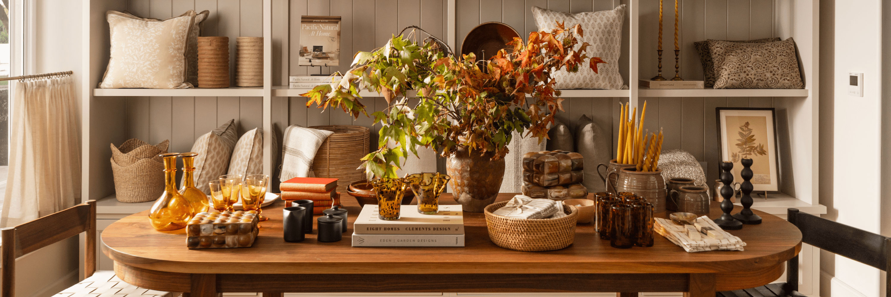 Styled wooden table with fall branches, amber glassware, books, candles, and baskets in a neutral shop display