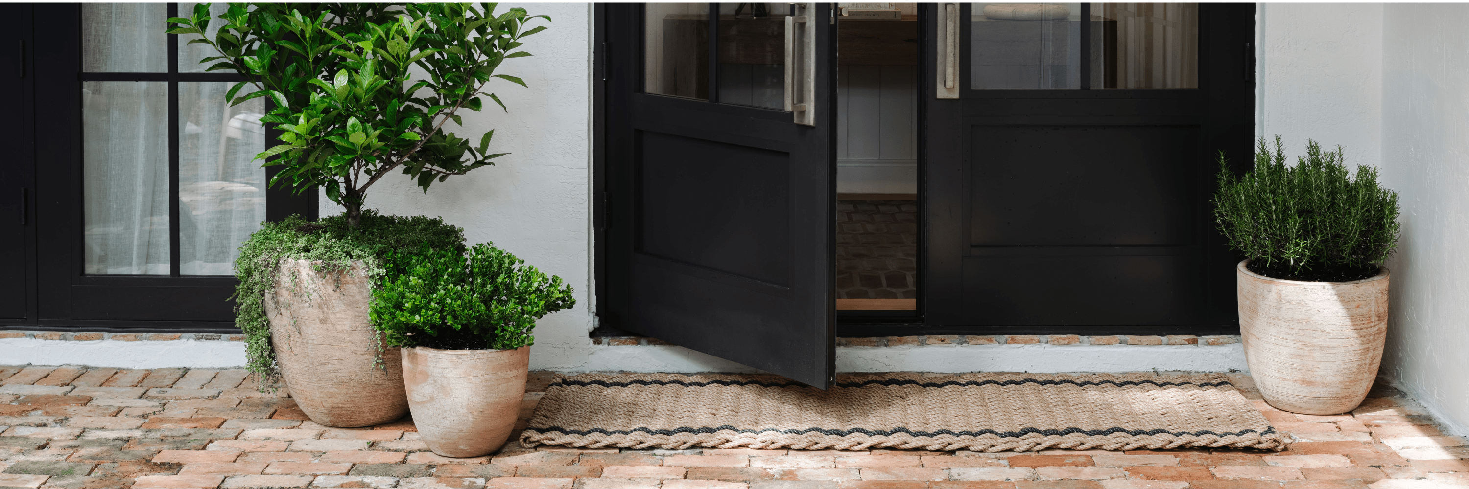 Outdoor entry with textured planters, lush greenery, and a woven doormat on brick flooring