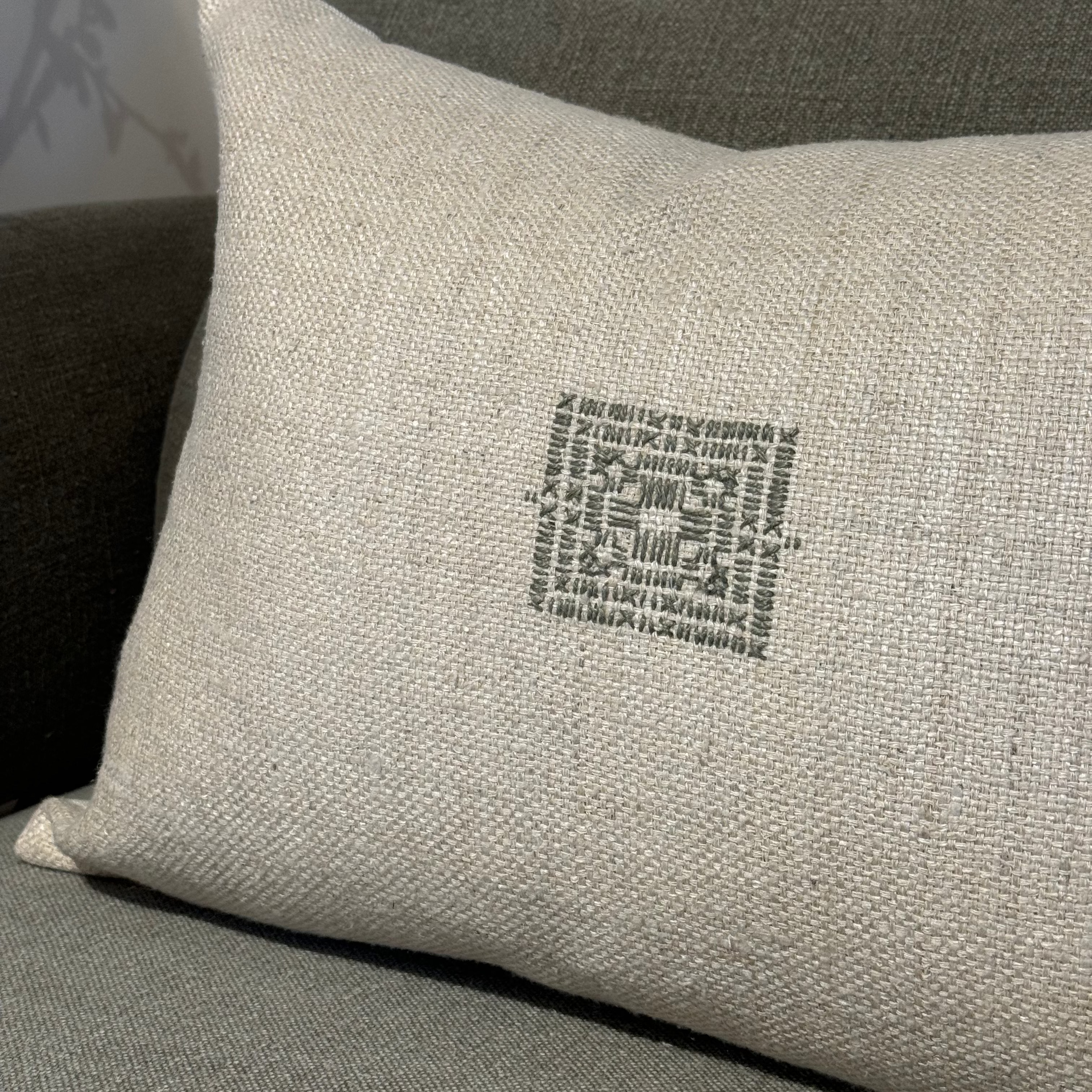 A beige square-shaped linen lumbar pillow with an embroidered geometric pattern.,