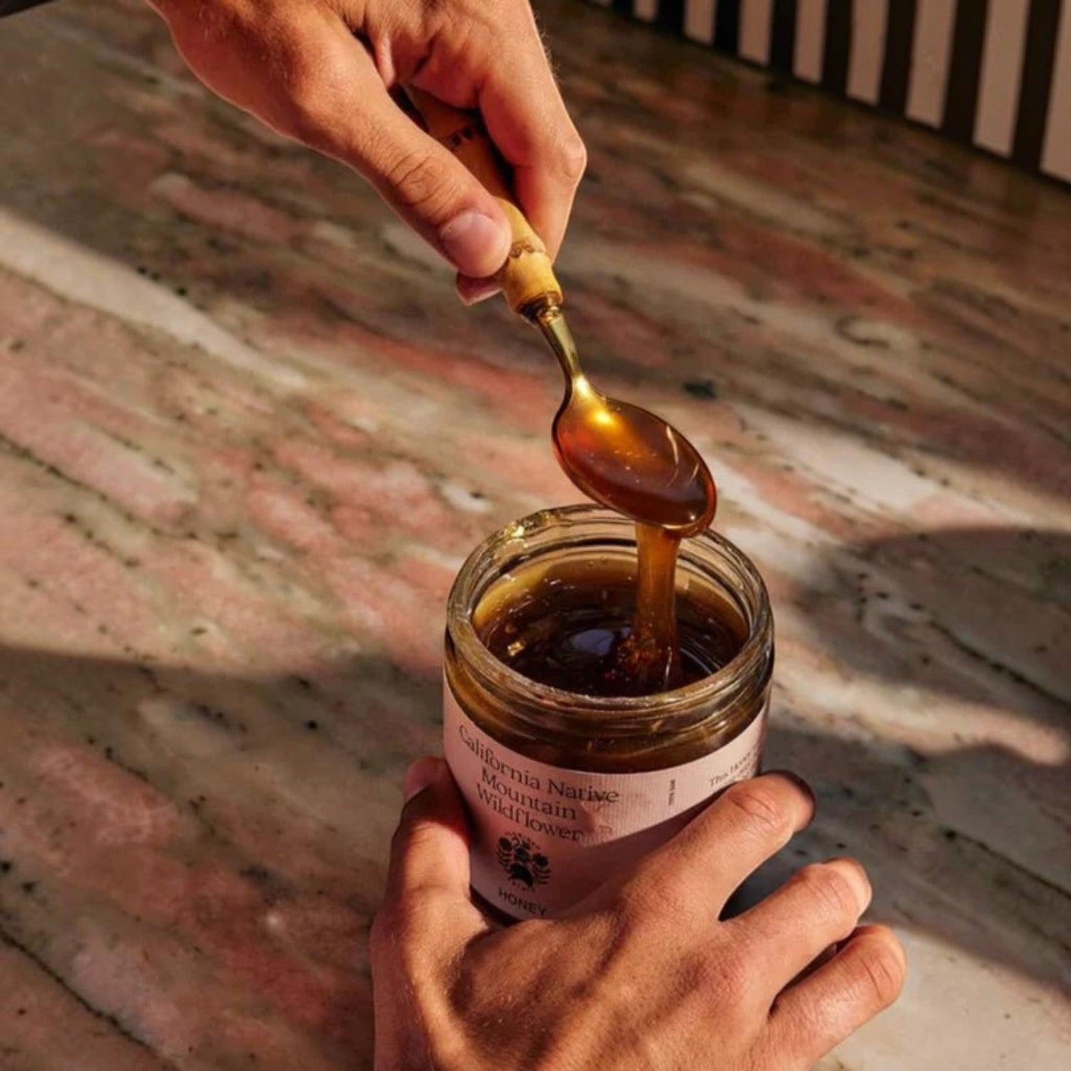 Hands cradle the Flamingo Estate California Mountain Wildflower Honey jar as a brass spoon drizzles thick golden honey back inside.