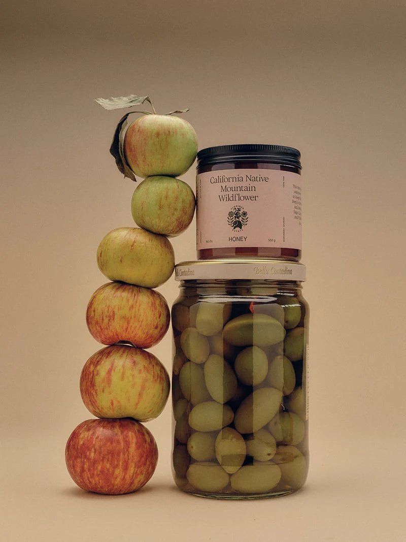 A Flamingo Estate California Mountain Wildflower Honey jar balances atop a jar of green olives next to a totem-stack of apples.