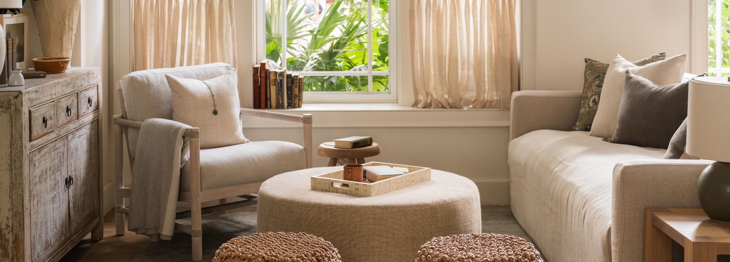 Warm living room interior with linen sofa, woven round ottoman, distressed wood sideboard, and tropical garden views designed by Calimia Home in Coral Gables