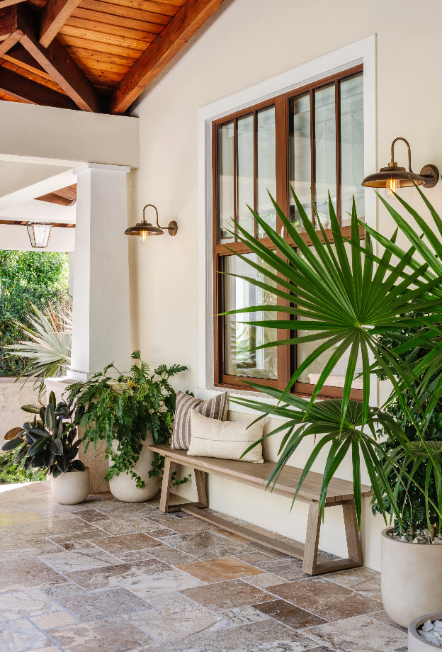 Front porch with woven bench, fan palms, and brass sconces under a warm wood ceiling
