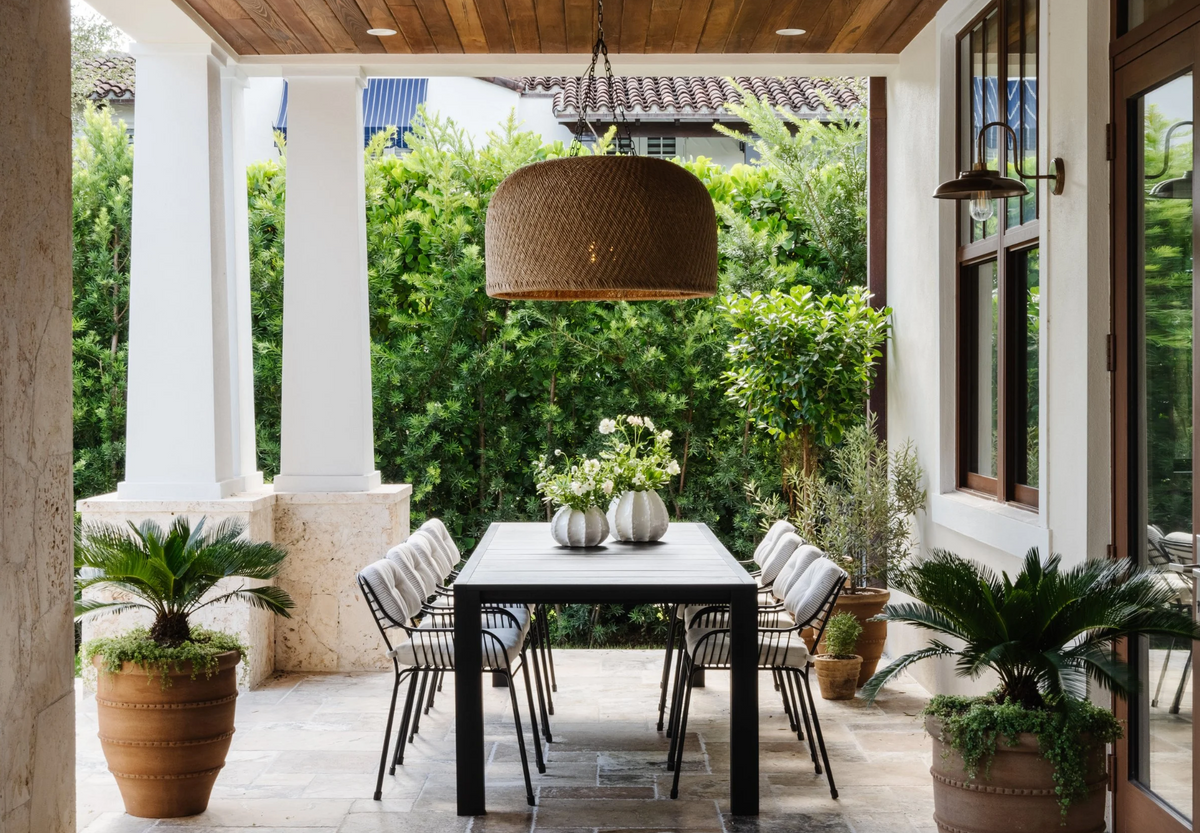 Outdoor dining table under an oversized rattan pendant surrounded by lush greenery