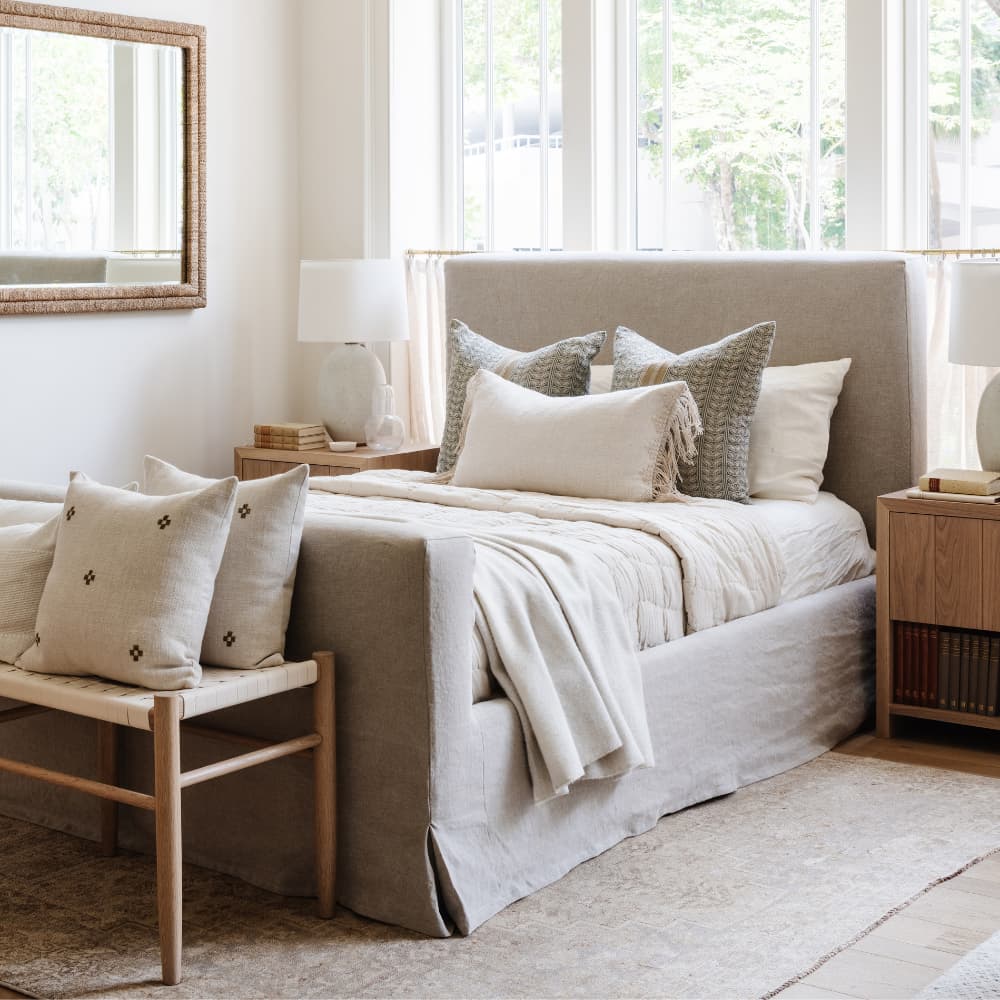 Primary bedroom with linen upholstered slipcovered bed, oak nightstands, woven bench, and layered neutral pillows designed by Calimia Home in Miami