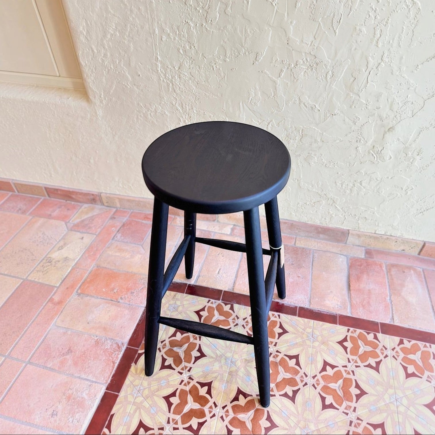 Calimia Home Omah Stool in black stained wood with a round seat and tapered legs on a tiled floor