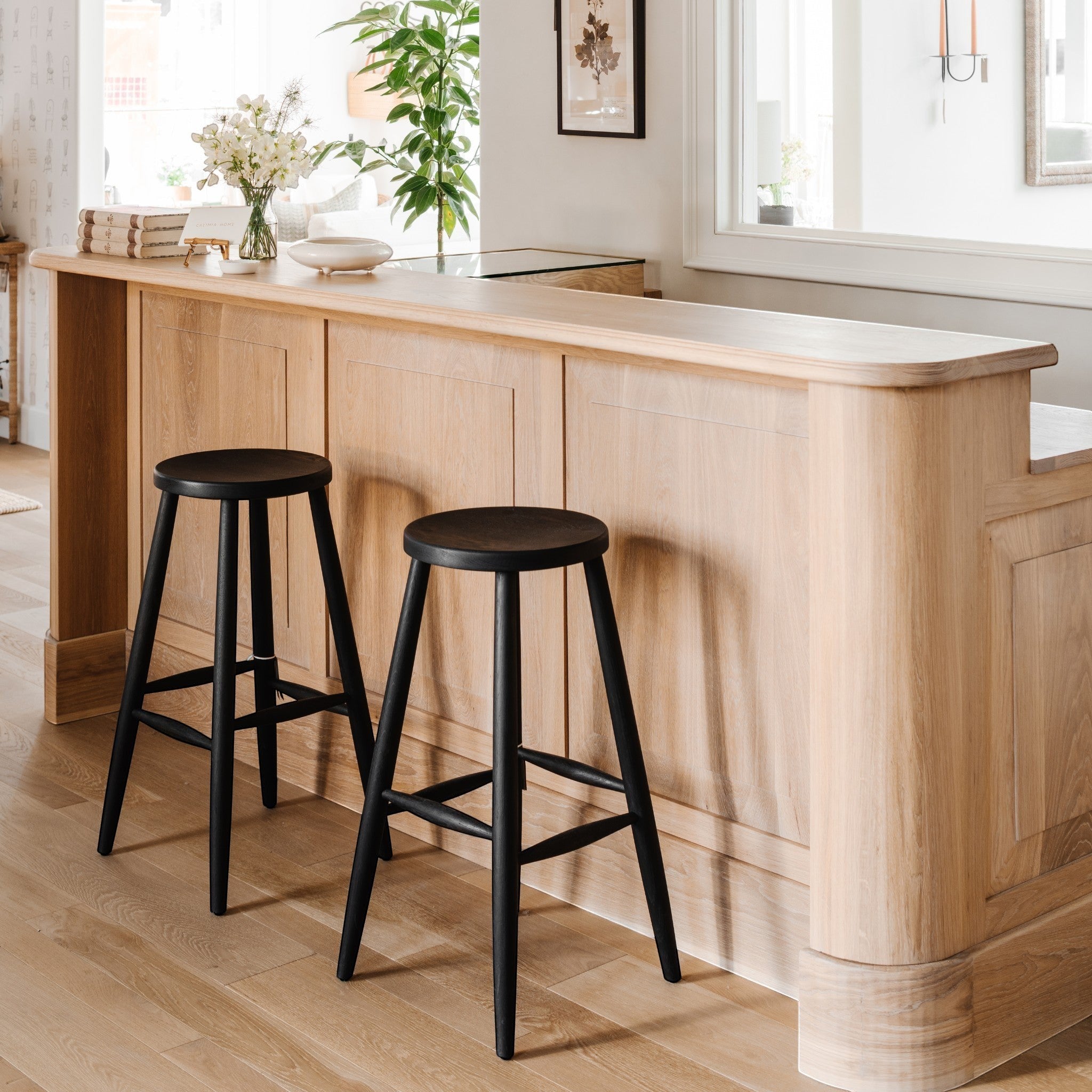 Two Calimia Home Omah stools with round black wooden seats and tapered legs positioned at a light oak breakfast bar.