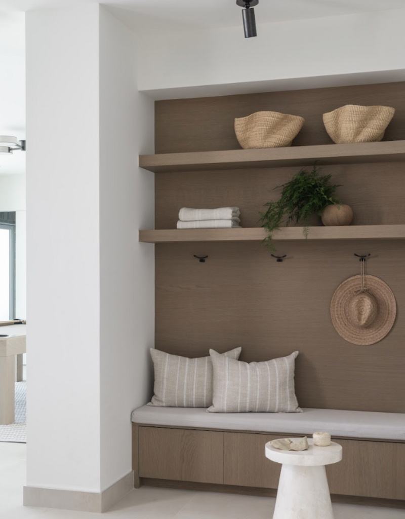 Built-in mudroom bench with striped pillows, oak shelves, woven baskets, and a sun hat