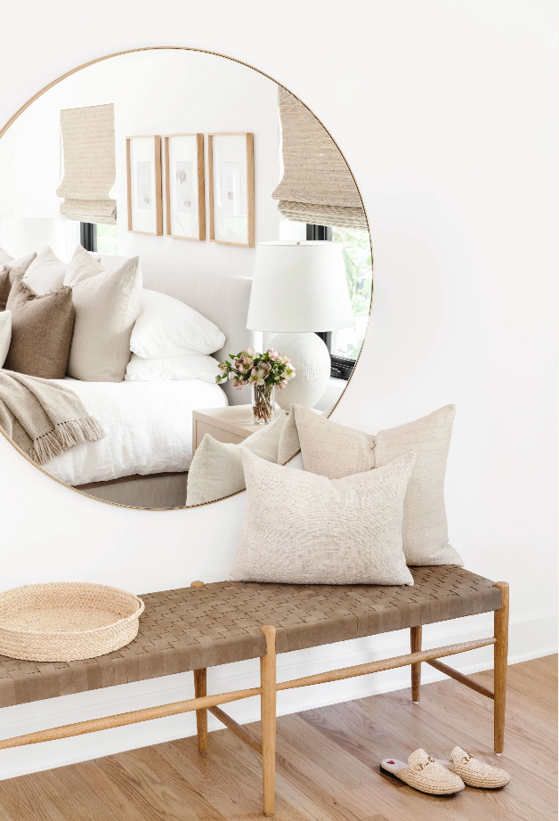 Round mirror above a woven leather bench stacked with linen pillows
