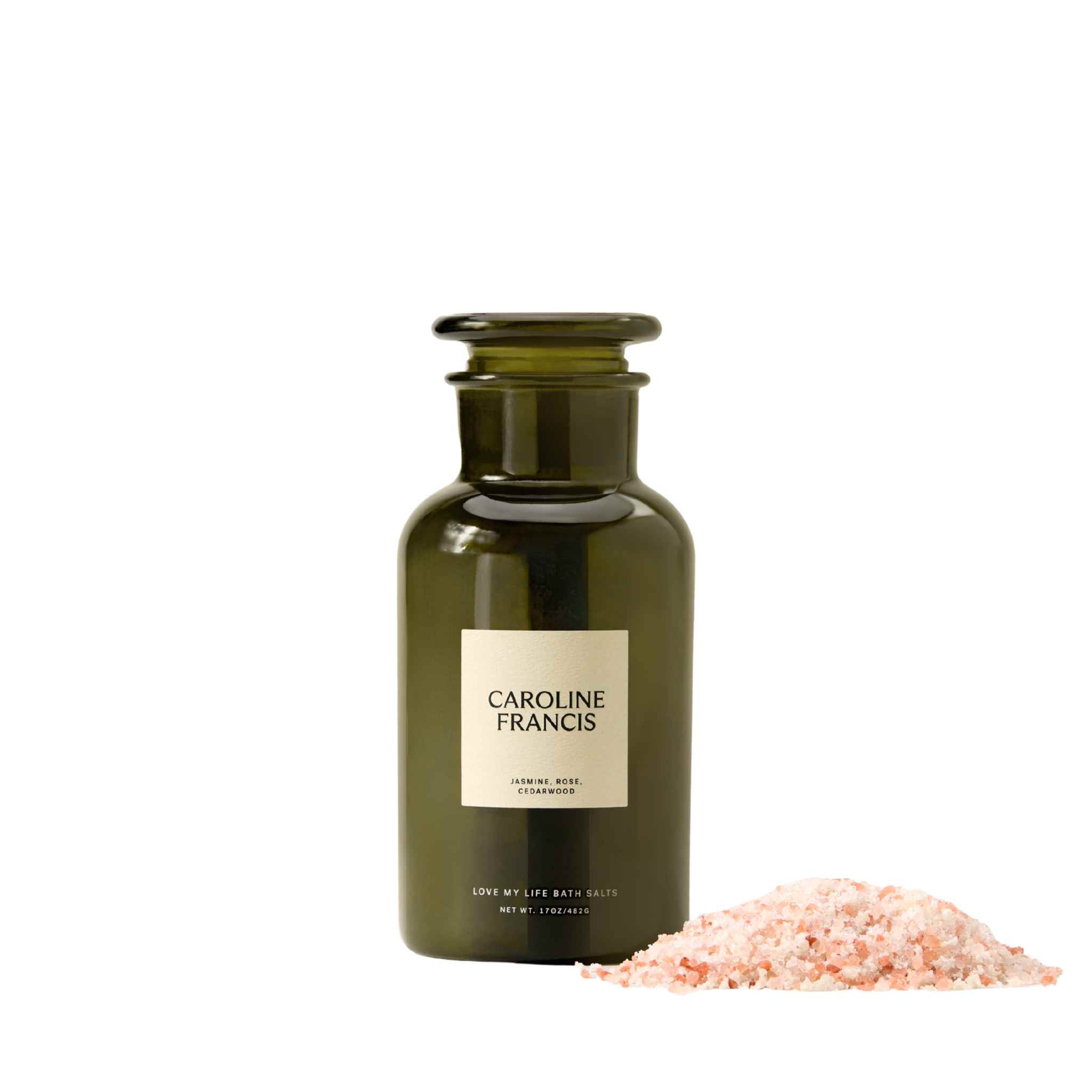 Caroline Francis Love My Life Bath Salts in a green glass apothecary bottle next to a loose pile of pink Himalayan bath salts.