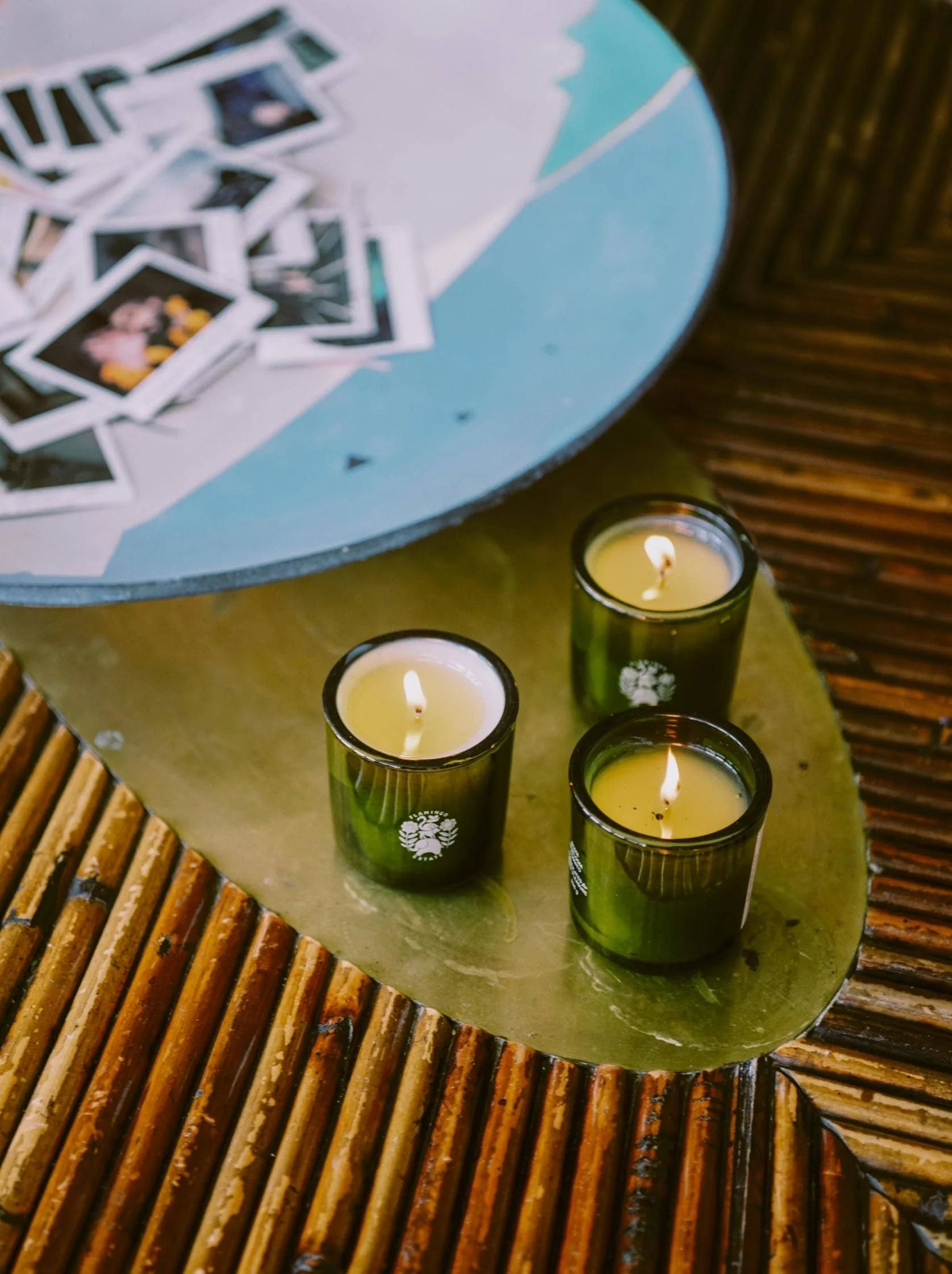 Three lit Flamingo Estate Douglas Fir & Ancient Vetiver votive candles glow on a brass tray atop a bamboo surface, scattered Polaroid photos curling at the edge.