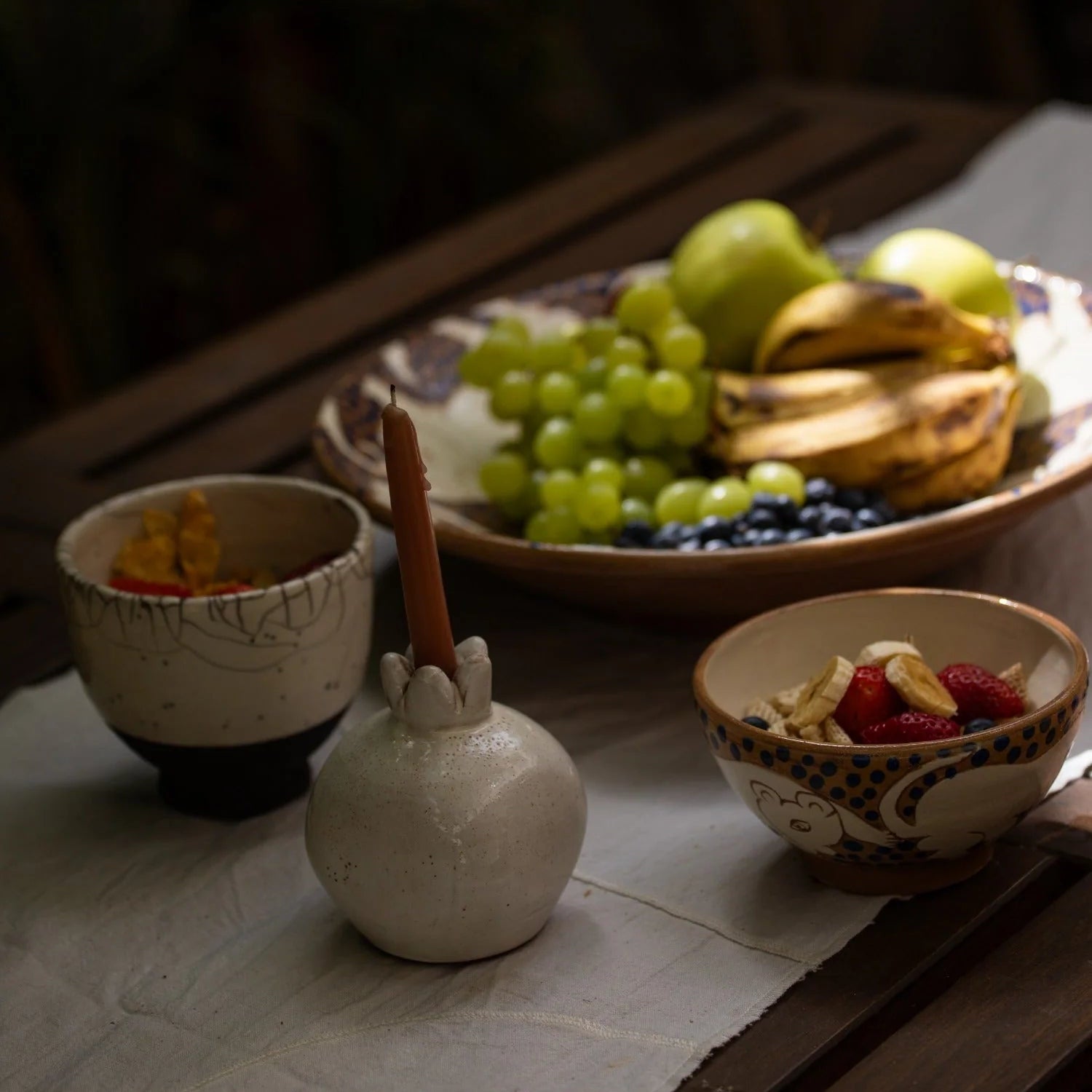 White ceramic pomegranate vase shown styled with a candle one a wooden table with fruit.