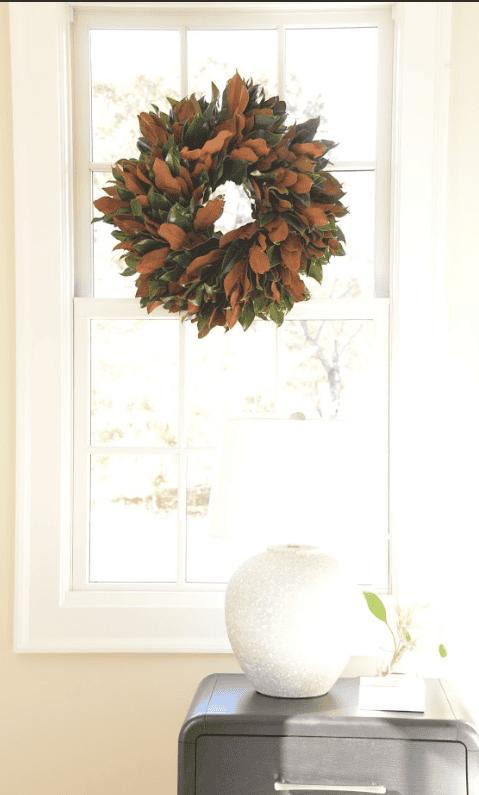 Fresh magnolia wreath hung in a sunlit window above a white ceramic vase