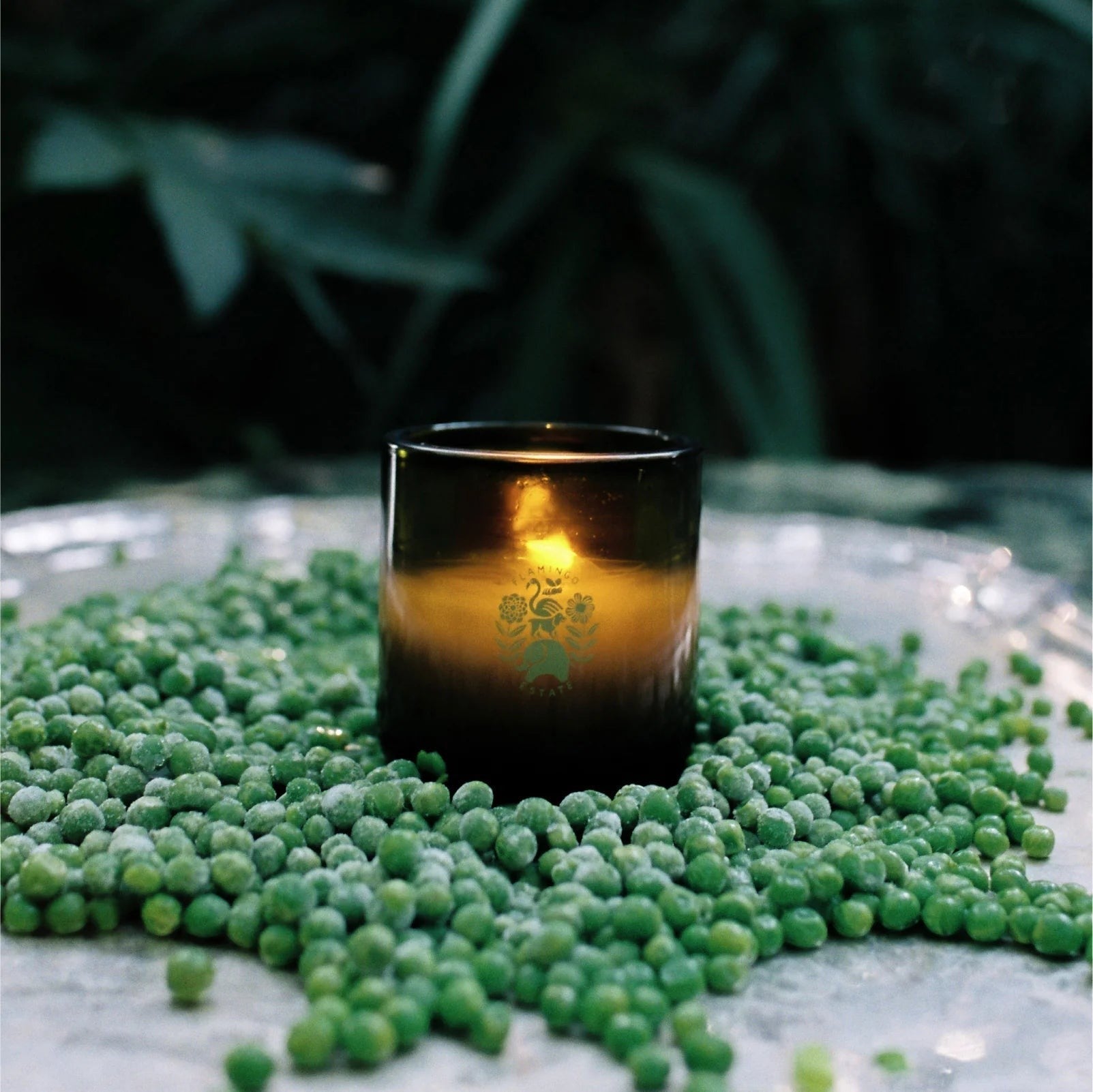 Flamingo Estate Green Goddess scented candle in a deep olive glass jar, glowing on a bed of fresh green peas.