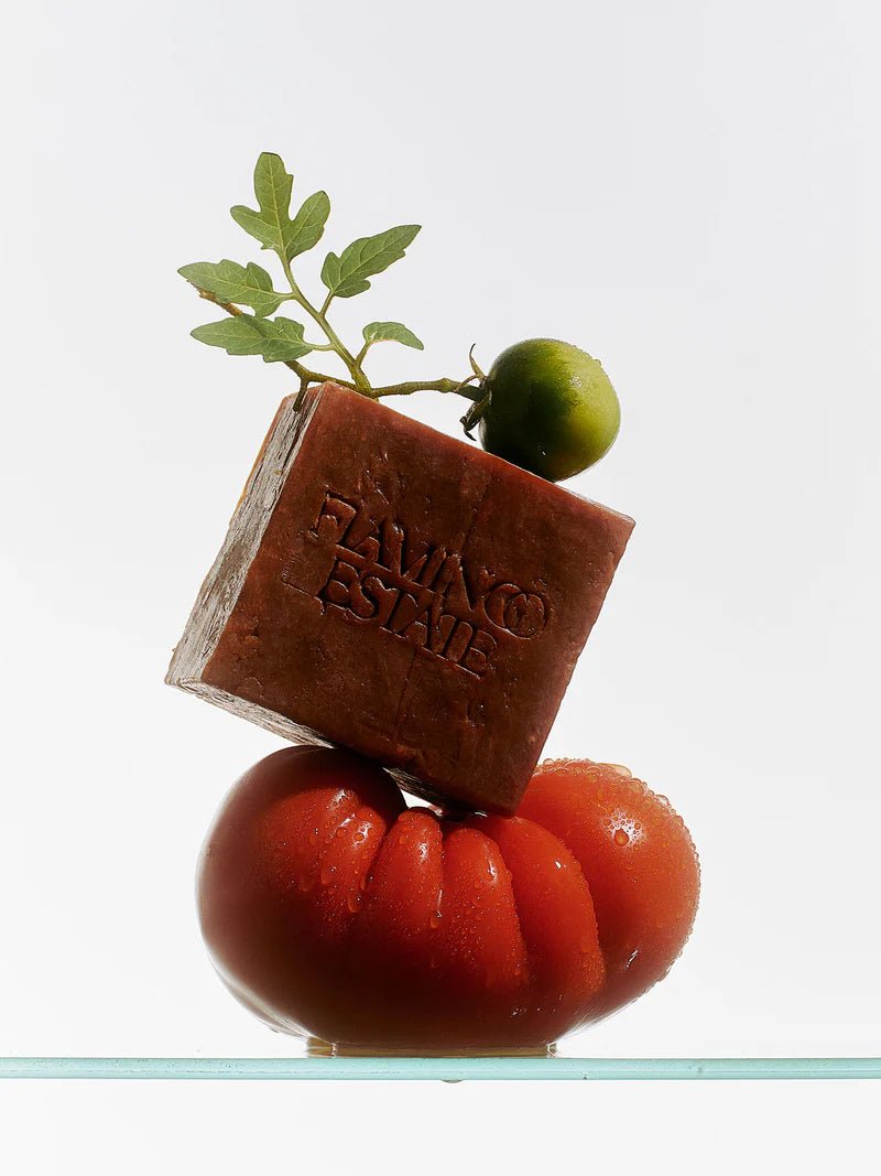 The brick-red Flamingo Estate Heirloom Tomato Soap Bar balances atop a glossy ripe red heirloom tomato, garnished with a small green tomato and leafy stem.