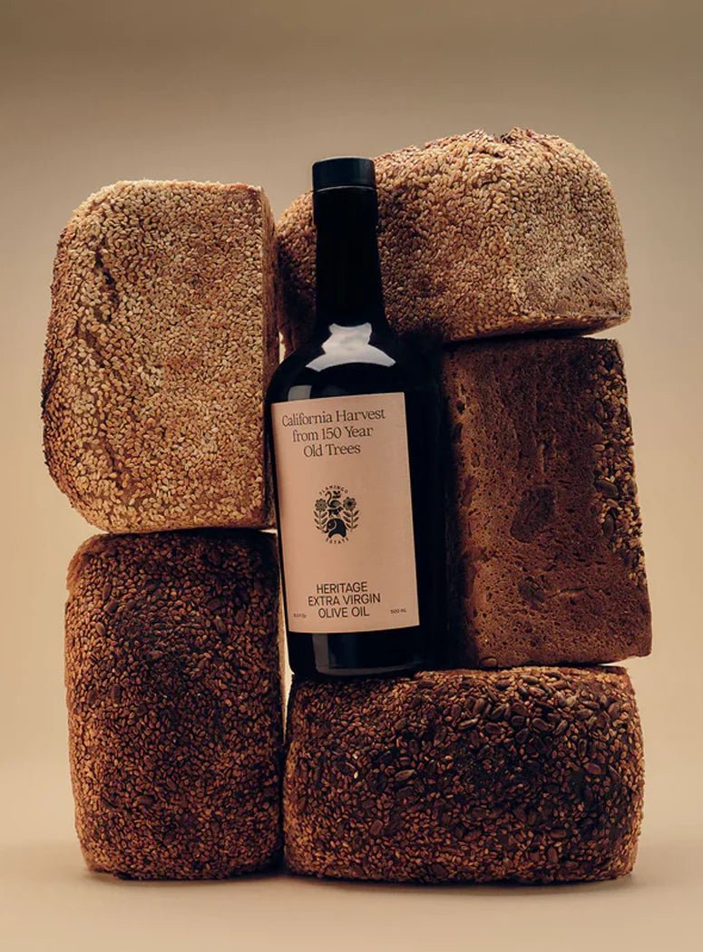 A Flamingo Estate Heritage olive oil bottle nestles between sculptural loaves of seeded brown bread stacked against a warm sand-toned backdrop.