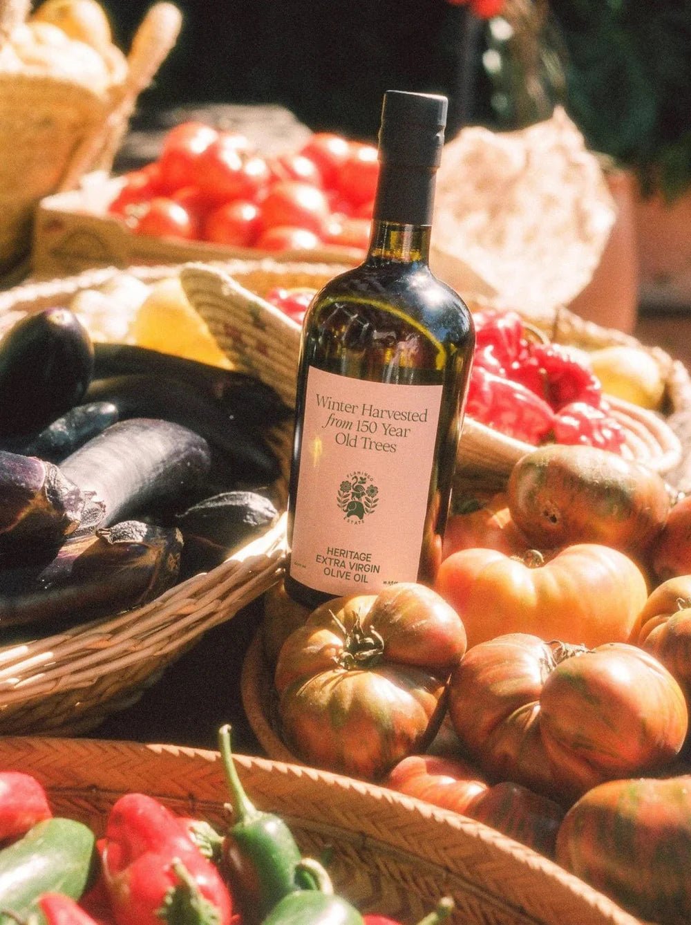 A Flamingo Estate olive oil bottle anchors a sunlit market spread of heirloom tomatoes, eggplants, peppers, and woven baskets.