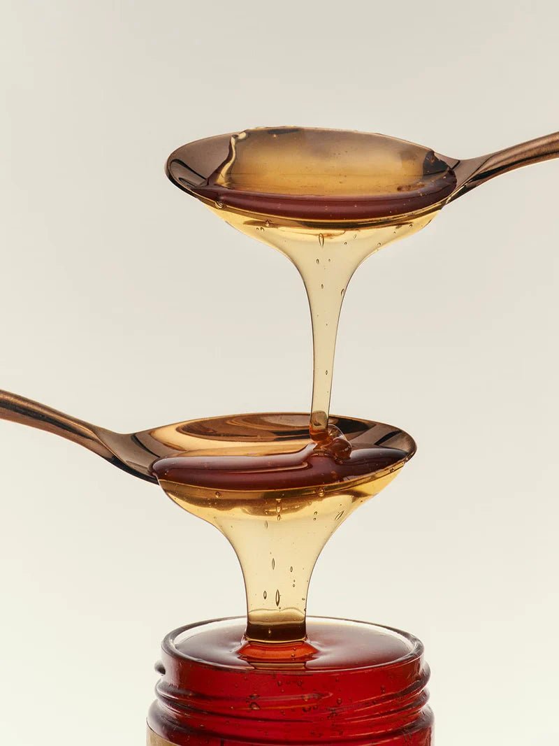 Two spoons cascade Flamingo Estate California wildflower honey, pale gold dripping into darker amber and into the open jar below.