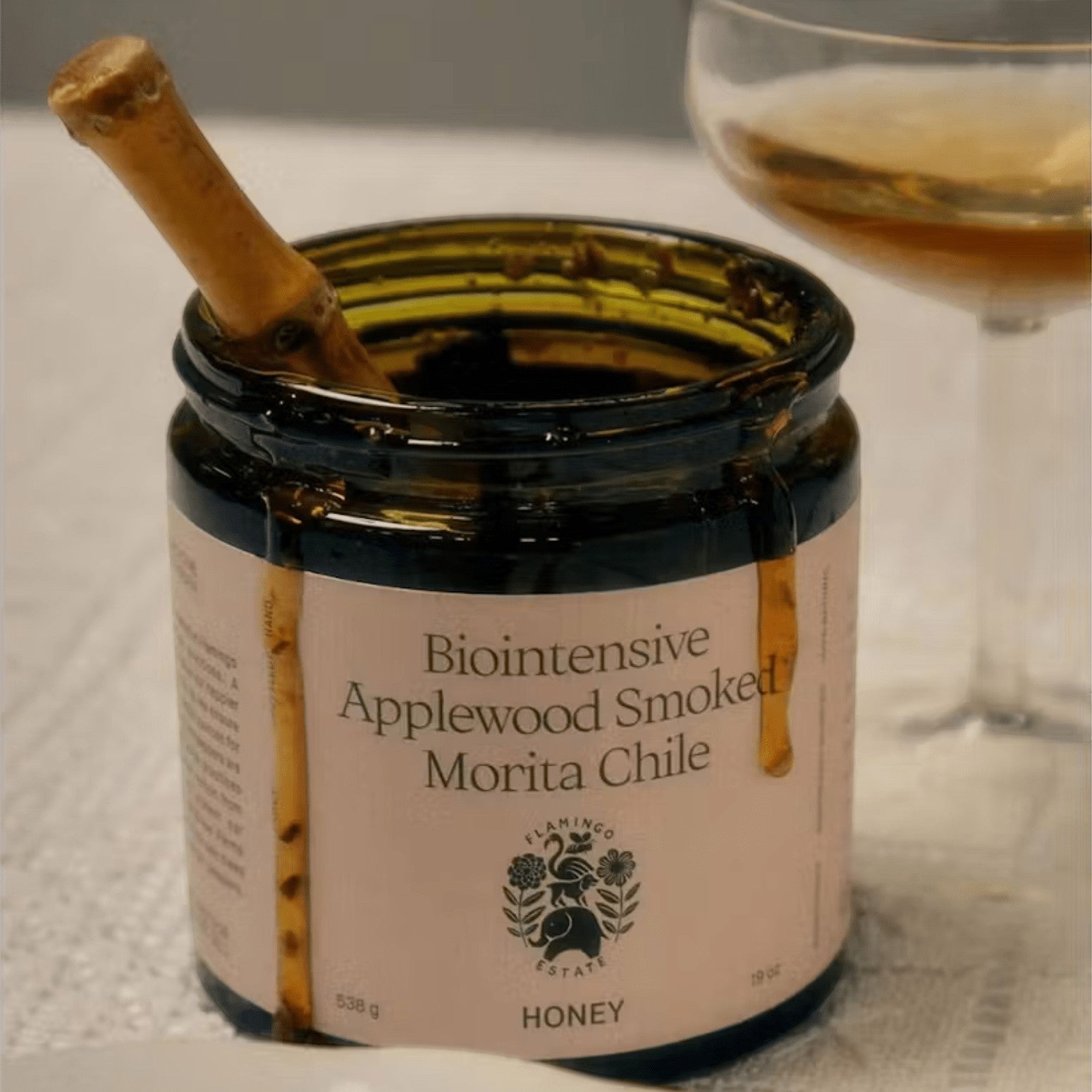 An open Flamingo Estate Applewood Smoked Morita Chile Honey jar with a wooden dipper inside, a coupe of amber wine alongside on linen.