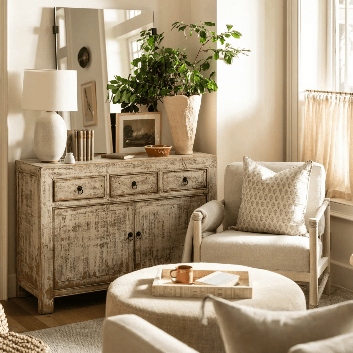 A styled living room interior featuring a large woven dome pendant light hanging over a distressed wood credenza, with a cream armchair, round ottoman, and potted green plants in a bright, sunlit room.