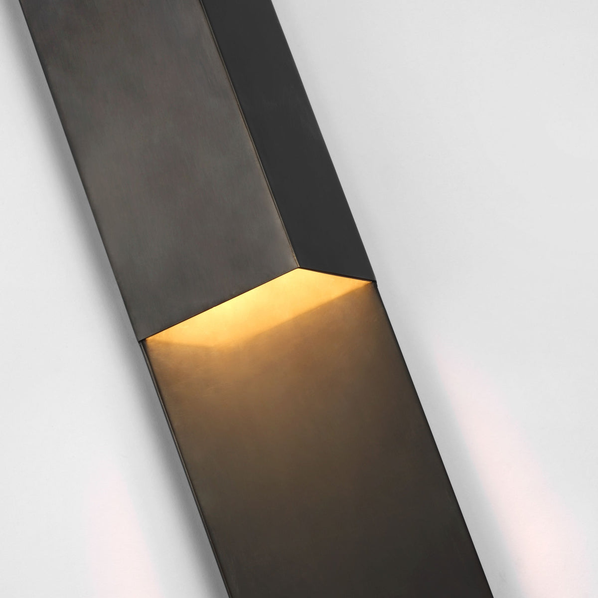 Close-up of the Rega sconce's folded bronze plane, warm LED light spilling across the lower panel