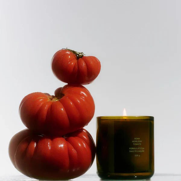 A lit Roma Heirloom Tomato Candle from Calimia Home Shop sits beside a stacked tower of three glossy red heirloom tomatoes against a bright white background.