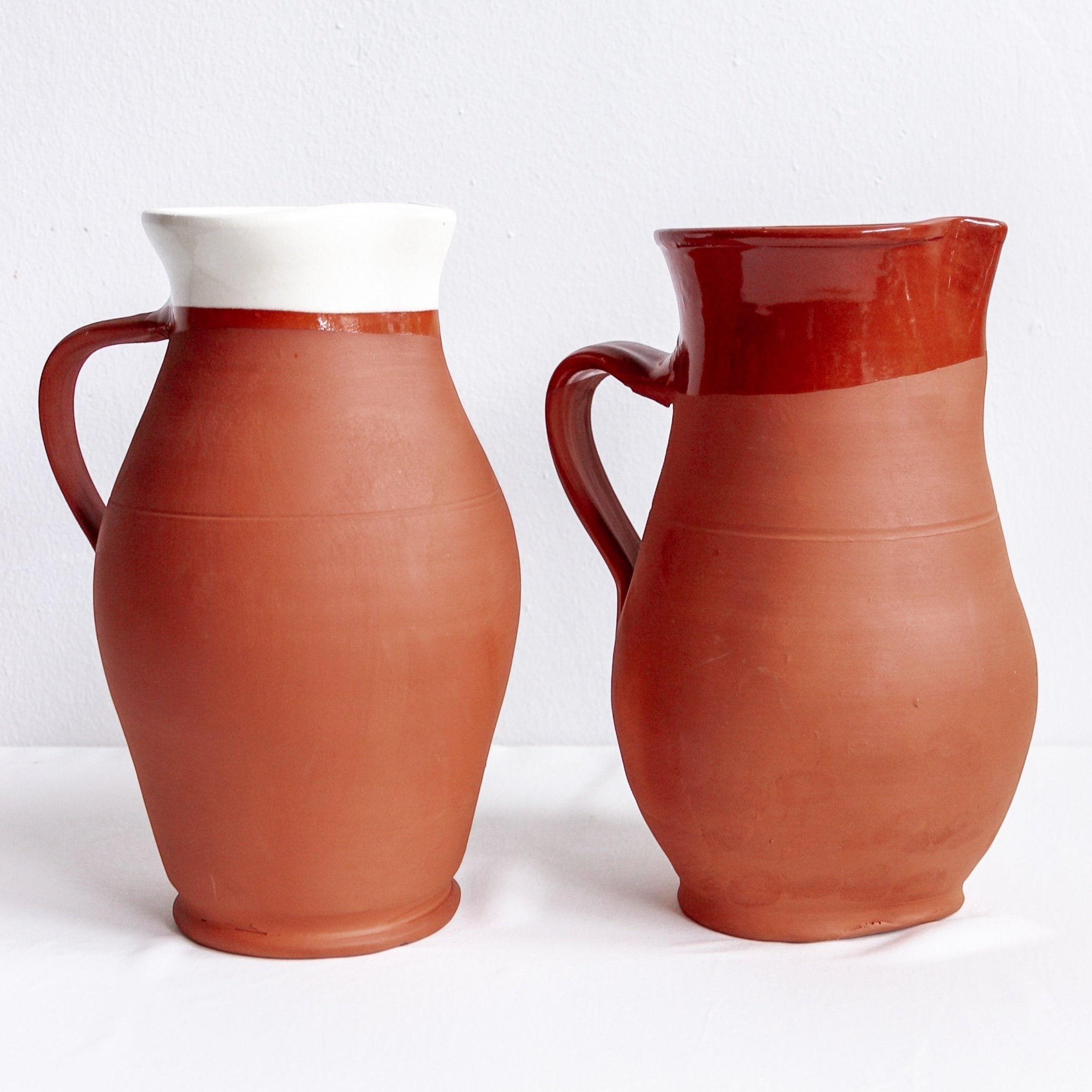 Two handmade Calimia Home terracotta pitchers with white and dark red glazed rims against a plain white background.