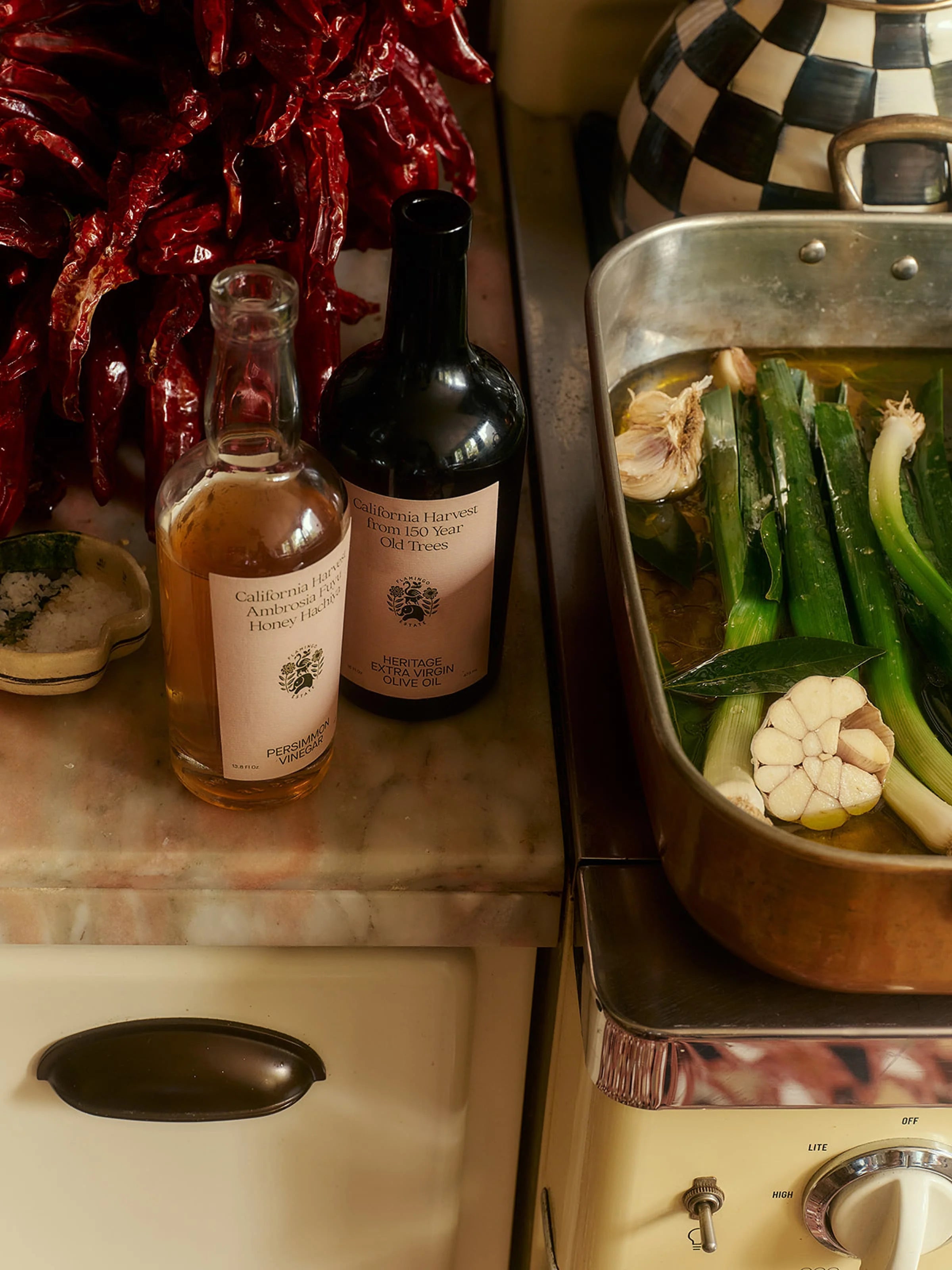 Flamingo Estate Persimmon Vinegar stands beside Heritage olive oil on a vintage marble counter with leeks, garlic, and dried chiles overhead.