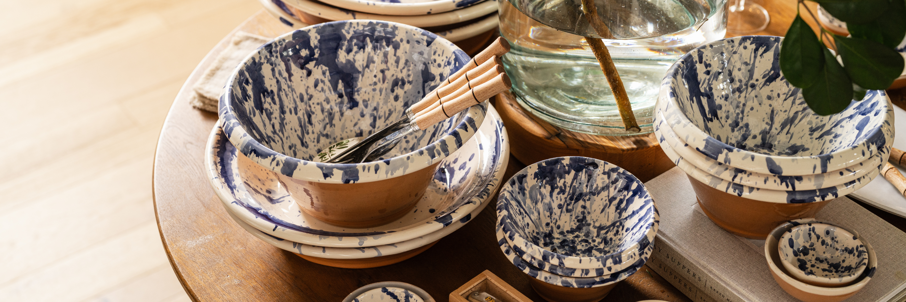 Round wooden dining table set with neutral dinnerware, linen napkins, and wildflowers in a glass pitcher. Soft light filters through linen curtains behind a cushioned bench at Calimia Home Shop.