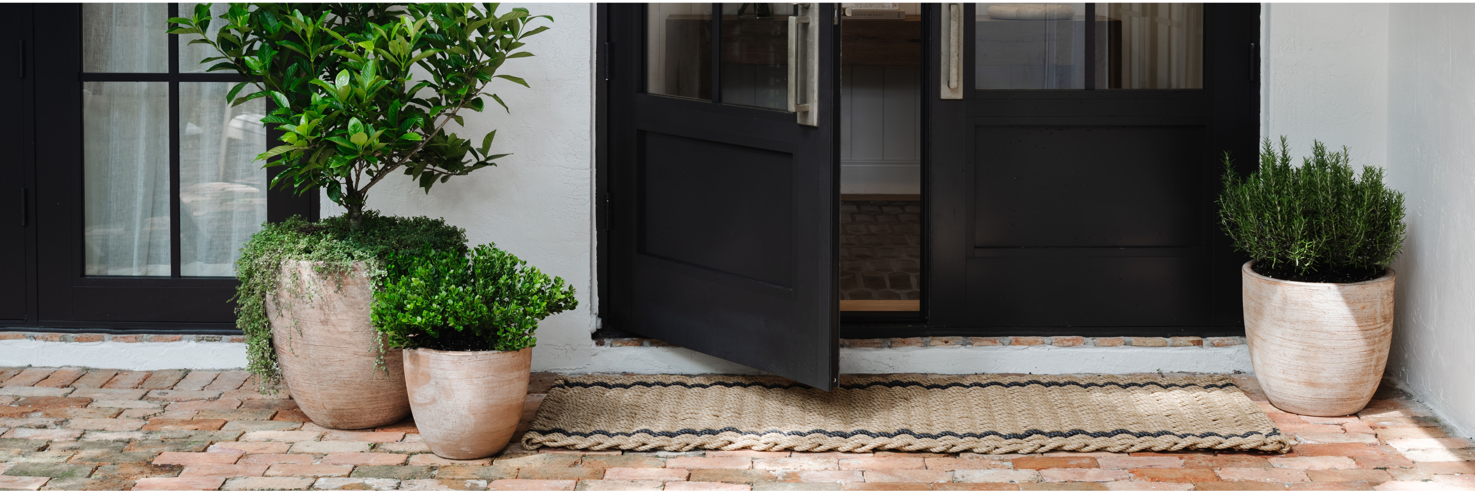 Outdoor entry with textured planters, lush greenery, and a woven doormat on brick flooring