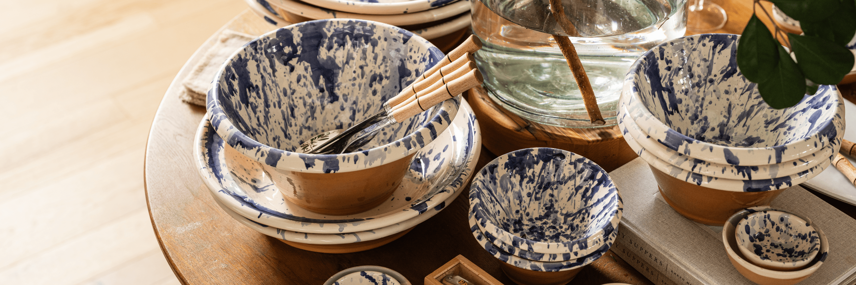 Round wooden dining table set with neutral dinnerware, linen napkins, and wildflowers in a glass pitcher. Soft light filters through linen curtains behind a cushioned bench at Calimia Home Shop.