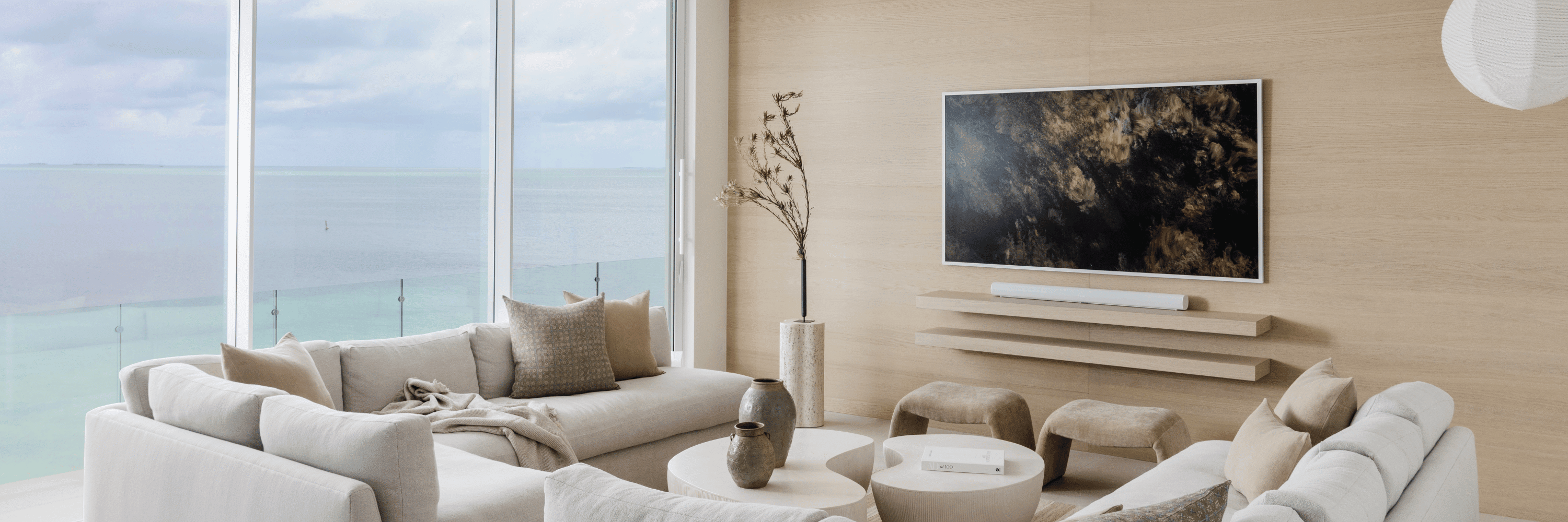 A sun-filled living room in soft neutrals — linen sectional, travertine coffee tables, woven stools — looking out through floor-to-ceiling windows onto a pale turquoise ocean