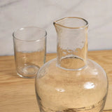 Be Home clear glass bedside carafe and matching tumbler with a textured pebble-like finish on a wooden surface.
