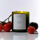 Flamingo Estate Heirloom Tomato scented soy wax candle in a green glass jar surrounded by fresh red tomatoes.