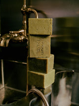Four green Flamingo Estate Rosemary & Parsley Soap Bricks stacked on a brass bathtub faucet with a shower hose.