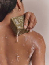 A hand lathers the green Flamingo Estate Rosemary & Parsley Soap Brick across a bare, sudsy shoulder.