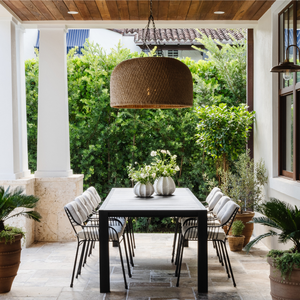 Outdoor dining area with a table and chairs under a large wicker pendant light, surrounded by greenery.