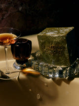 A lathered Flamingo Estate Rosemary & Parsley Soap Brick on a glass soap dish beside a cocktail glass on a bath ledge.