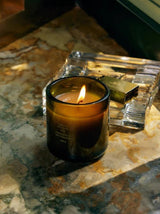 A lit Adriatic Muscatel Sage Candle rests on a veined marble ledge beside a cut-crystal box and brass match holder, its single flame glowing in the moody half-light.