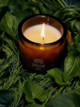 A lit Ancient Agrigento Olive Tree Candle nestles in a lush wreath of basil, mint and cypress sprigs, weaving together the green herbal notes of its fragrance.