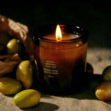 A brown glass Ancient Agrigento Olive Tree Candle from Flamingo Estate burns on a linen cloth, encircled by fresh green olives that mirror its Mediterranean grove scent.
