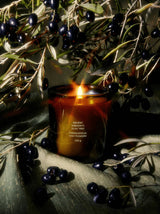 A glowing Ancient Agrigento Olive Tree Candle sits on draped olive-green fabric beneath hanging olive branches heavy with dark ripe olives — a sun-warmed Sicilian still life.