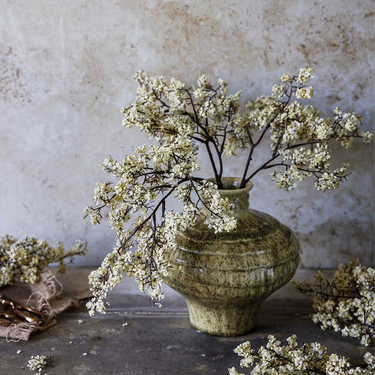 Artificial Sedge Winter Dew stems by Abigail Ahern arranged in a green ceramic vase against a plaster wall