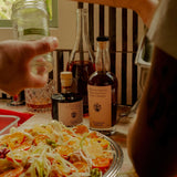 Flamingo Estate Balsamic Vinegar joins the Persimmon Vinegar at a citrus salad table, a hand reaching past a striped chair in warm light.