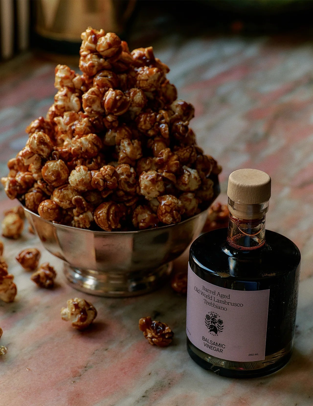 A Flamingo Estate Barrel Aged Balsamic Vinegar bottle stands on marble beside a silver footed bowl piled high with caramel-glazed popcorn.