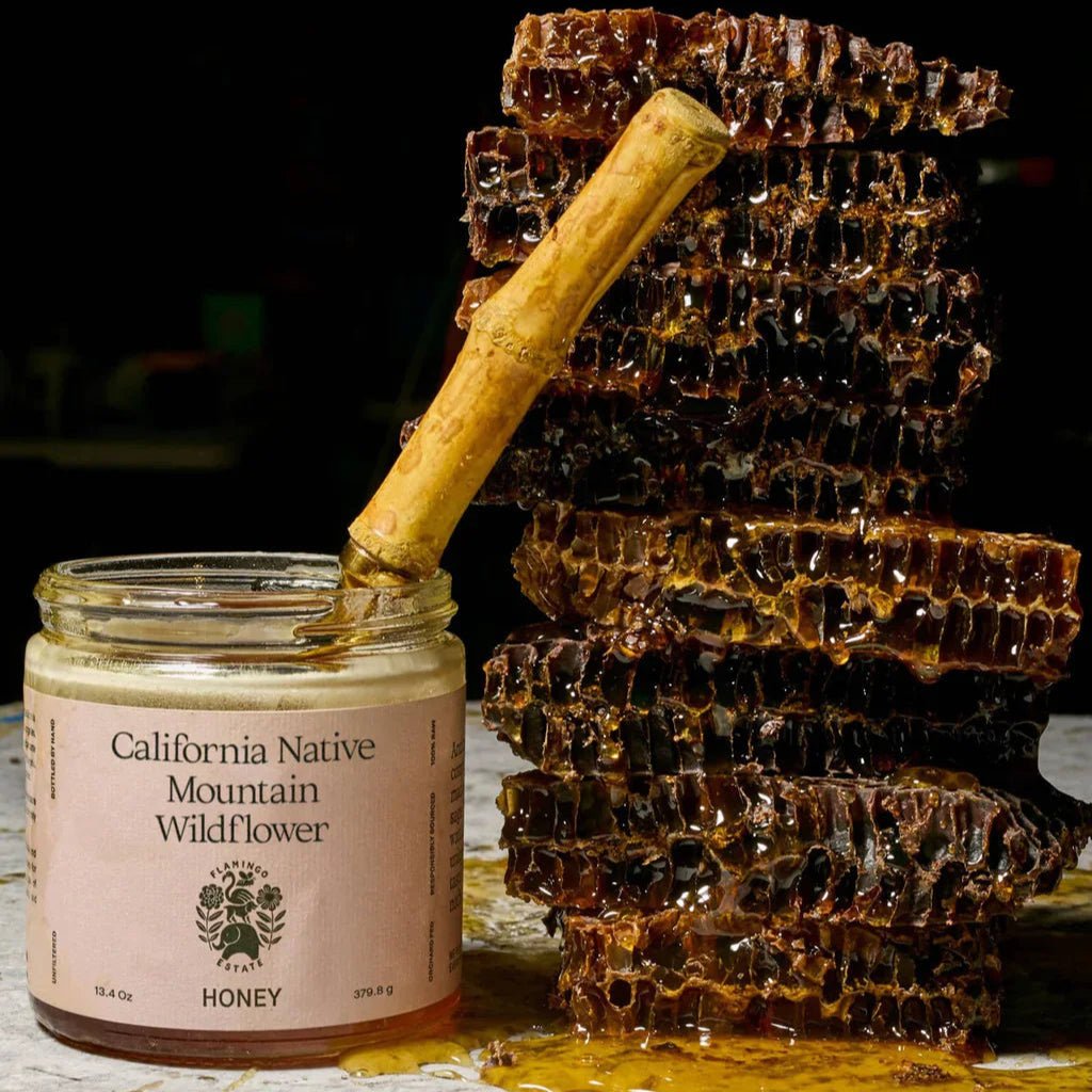 An open Flamingo Estate California Mountain Wildflower Honey jar with a wooden dipper sits beside a tall stack of dripping raw honeycomb.