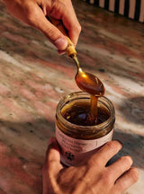 Hands cradle the Flamingo Estate California Mountain Wildflower Honey jar as a brass spoon drizzles thick golden honey back inside.